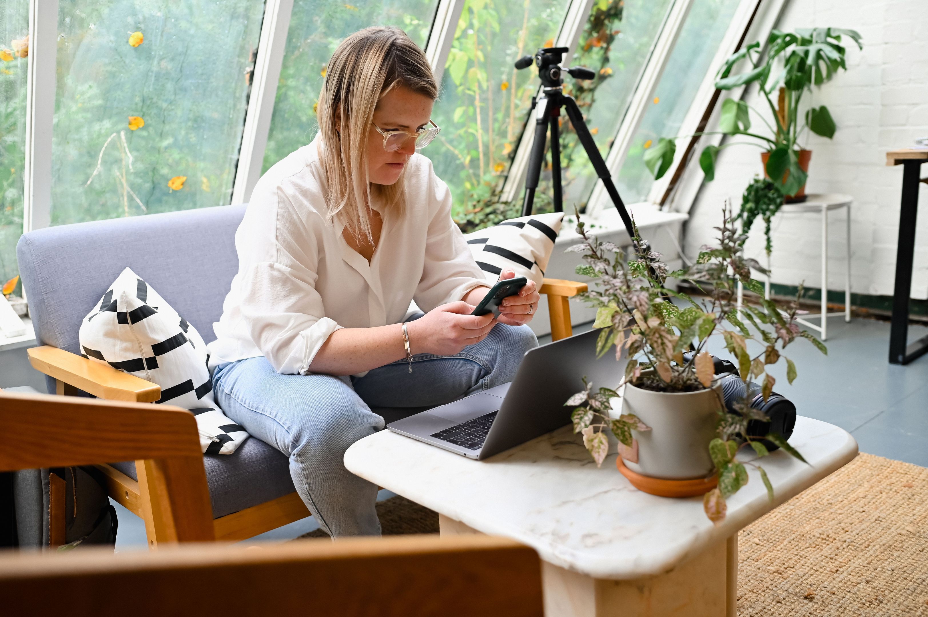 A photographer sits on a couch in front of their laptop, looking at their mobile device as files are backed up to the cloud.